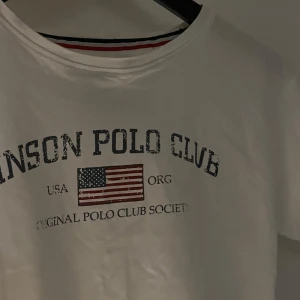 Vit t-shirt  - Vit t-shirt från Vinson Polo Club med tryck av amerikansk flagga och texten 'Original Polo Club Society'. 