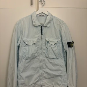 Ljusblå overshirt från Stone Island - Säljer en stilren ljusblå overshirt från Stone Island med dragkedja och två bröstfickor. Den har långa ärmar och en klassisk krage. Perfekt för lager-på-lager-stil.