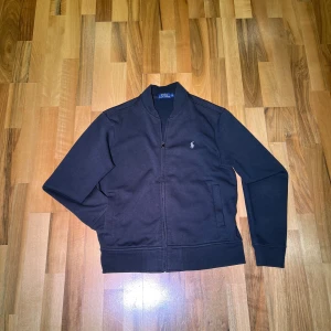 Svart cardigan/tröja från Polo Ralph Lauren M - Snygg svart cardigan/tröja från Polo Ralph Lauren med dragkedja och broderad logga på bröstet. Perfekt för en stilren look. Plaggen har långa ärmar och är perfekt för kylig väder. Äkta. M. Kan se lite urtvättad ut