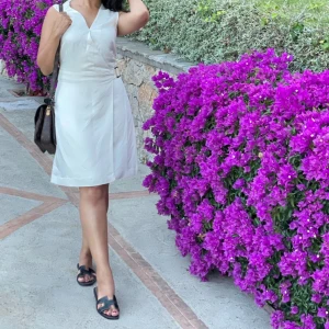 Vit omlottklänning med bälte - Snygg vit omlottklänning med ett stilrent bälte i midjan. Klänningen har en v-ringad design och är ärmlös, vilket gör den perfekt för varmare dagar. Den har en dragkedja i sidan för enkel på- och avtagning.