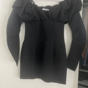 Svart klänning med volangdetaljer - Elegant svart klänning med volangdetaljer vid axlarna och långa ärmar. Perfekt för en stilren look.