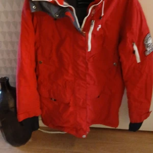 Röd Peak Performance jacka i stl L - Snygg röd jacka från Peak Performance med dragkedja och huva. Jackan har den äldre broderade loggan på ärmen och flera praktiska fickor. Perfekt för kyligare dagar.