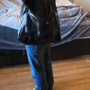 Svart axelväska i skinn - Snygg och rymlig svart axelväska i skinn med långa remmar. Perfekt för att bära med sig allt man behöver under dagen. Enkel och stilren design som passar till många olika outfits.