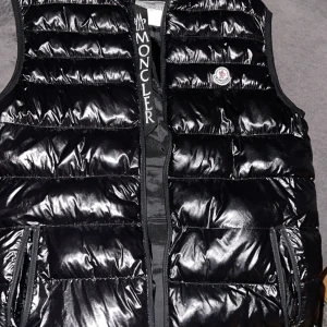Svart dunväst från Moncler - Snygg svart dunväst från Moncler med glansig finish och dragkedja framtill. Västen har Moncler-logga på bröstet och är perfekt för kyligare dagar. Den är quiltad för extra värme och stil.