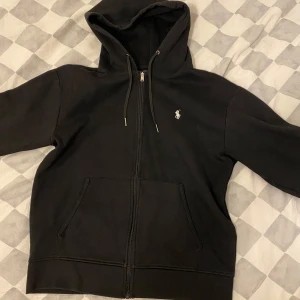 Svart hoodie från Polo Ralph Lauren - Säljer en klassisk svart hoodie från Polo Ralph Lauren med dragkedja och huva. Den har en liten vit logotyp på bröstet och praktiska fickor framtill. Perfekt för en avslappnad stil. 