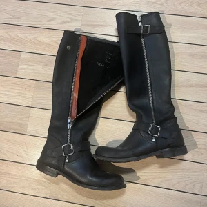 Svarta höga stövlar med dragkedja - Snygga svarta höga läder stövlar i skinn med dragkedja och spänne. Från Storbritannien. Perfekta för höst och vinter. Robust sula för bra grepp. Sparsamt använda. Nypris 1500kr