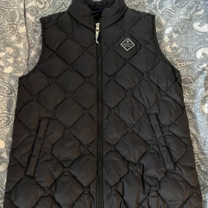 Svart quiltad väst från Gant - Snygg svart quiltad väst från Gant med dragkedja och en liten logotyp på bröstet. Perfekt för lager-på-lager under kyligare dagar. Passar ungdomar i åldern 13-14 år.