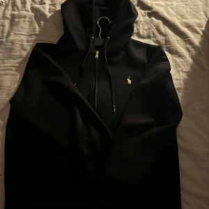 Svart hoodie från Ralph Lauren - Snygg svart hoodie från Ralph Lauren med dragkedja och klassisk logga på bröstet. Perfekt för en avslappnad stil. Den har en huva med snörning och är tillverkad i mjukt material. 🐎