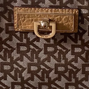 Brun mönstrad axelväska från DKNY - Snygg brun axelväska från DKNY med ett monogrammönster och gulddetaljer. Väskan har en dragkedja och en elegant kedjerem. Perfekt för att ge din outfit en lyxig touch.