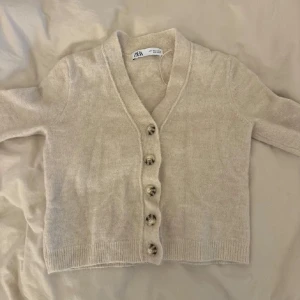 Beige kofta från Zara - Säljer en stilren beige kofta från Zara med knappar framtill. Koftan har en v-ringad design och är tillverkad i ett mjukt material som passar perfekt för kyligare dagar.