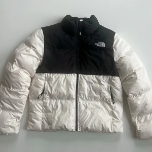 Vit och svart dunjacka från The North Face - 1996 Retro Nuptse från The North Face i storlek M. Jackan är vit med svarta detaljer och har en klassisk pufferstil. Perfekt för kalla vinterdagar med sin långa ärmar och dragkedja framtill. Den är i nyskick och redo för nya äventyr!