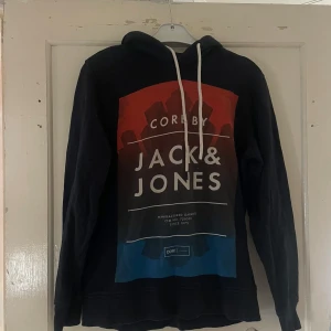 Svart hoodie från Jack & Jones - Säljer en snygg svart hoodie från Jack & Jones. Den har en stor logga på framsidan i rött och blått, vilket ger den en cool look. Perfekt för höst och vinter med sin mysiga huva och långa ärmar. Passar både till vardags och chillkvällar.