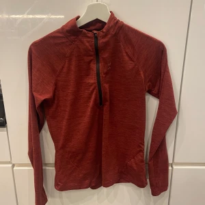 Röd långärmad tröja - Säljer en snygg röd långärmad träningströja. Perfekt för träning eller utomhusaktiviteter. Tröjan har en halv dragkedja framtill och en reflekterande detalj på ryggen. Materialet är lätt och andas bra. Märket har börjat ramla av lite på framsidan❤️
