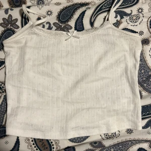 Vit ribbad linnetopp med spetsdetaljer - Säljer en söt vit ribbad linnetopp med smala axelband och spetsdetaljer längs halsringningen. Toppen har en liten rosett framtill och är perfekt för sommardagar. Den är i bra skick och superbekväm att ha på sig!