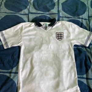 Retro England fotbollströja  - Säljer en klassisk vit retro England fotbollströja  som är i storlek S. Tröjan har korta ärmar med svarta och vita ränder vid ärmsluten och en svart krage med vita detaljer. Perfekt för fotbollsfans som gillar retrostil!
