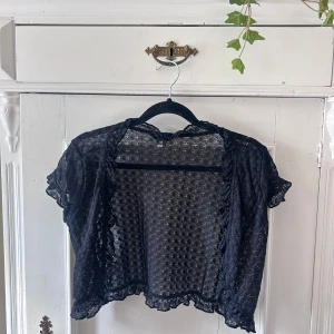 Svart spetsbolero i storlek XS - Säljer en superfin svart bolerotopp i spets. Den är kortärmad och har ett vackert mönster som ger en elegant touch. Perfekt för att piffa upp en outfit till fest eller en speciell kväll. Toppen är i nyskick och kommer från Polen.