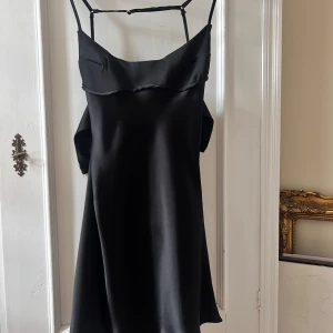 Svart axelbandslös klänning - Säljer en elegant svart axelbandslös klänning i satin. Den har en stilren design med smala axelband och en mjuk, glansig yta. Perfekt för festliga tillfällen eller en utekväll. Klänningen är i nyskick och ger en lyxig känsla.