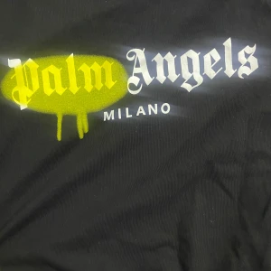Svart t-shirt från Palm Angels - Säljer en svart t-shirt från Palm Angels med ett coolt tryck på framsidan där det står 'Palm Angels Milano' i vitt och gult. T-shirten är i bomull och har en normal    passform. Perfekt för en avslappnad stil den är oxå helt äkta  Pris går diskutera