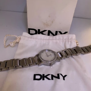 Silverfärgad klocka från DKNY - Säljer en elegant silverfärgad klocka från DKNY. Klockan har en stilren design med en glittrig urtavla och stenar runt boetten. Armbandet är i metall och har en klassisk länkstil. Perfekt för både vardag och fest!