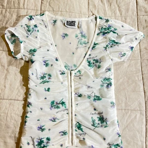 Blommig topp från Reclaimed Vintage - Säljer en superfin blommig topp från Reclaimed Vintage i storlek S. Den är vit med gröna och lila blommor och har en djup v-ringning. Toppen är kortärmad och har en söt knytning framtill. Perfekt för sommardagar eller en avslappnad vårlook!