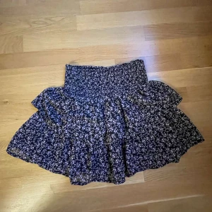 Blommig kjol från KappAhl - Säljer en superfin blommig kjol från KappAhl i storlek 146/152. Kjolen har en mörkblå bas med små vita blommor och en volangdetalj som ger den extra volym. Perfekt för vår och sommar!