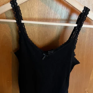 Svart linne från H&M Divided - Säljer ett svart linne från H&M Divided i storlek XS. Det är i nyskick och har en söt liten rosett framtill. Linnet har spetsdetaljer vid axelbanden och halsringningen, vilket ger det en extra touch av stil. Perfekt för både vardag och fest!