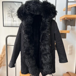 Svart parkajacka med pälsfoder - Säljer en supermysig svart parkajacka med fluffigt pälsfoder. Den har en stor huva med päls som håller dig varm under kalla dagar. Jackan är perfekt för vintern och har långa ärmar. Den är både snygg och praktisk, perfekt för att hålla stilen när det är kallt ute.