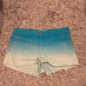 Blå ombre shorts från Hollister - Säljer ett par snygga blå ombre shorts från Hollister. De har en fräsch övergång från mörkblått till ljusblått och är prydda med en knapp och dragkedja framtill. Shortsen har fransiga kanter och klassiska bakfickor med en liten logga på framsidan. Perfekta för sommardagar! 🌊
