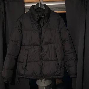 Svart pufferjacka - Säljer en svart pufferjacka med dragkedja och långa ärmar. Jackan har en hög krage och är perfekt för kyligare väder. Den quiltade designen ger extra värme och stil. Perfekt för vintern!