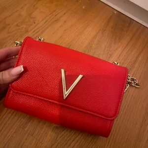 Röd axelväska med gulddetaljer - Snygg röd axelväska med en elegant guldfärgad kedja och ett stort V-emblem på framsidan. Väskan har en praktisk tryckknappsstängning och är perfekt för att bära dina viktigaste saker med stil.