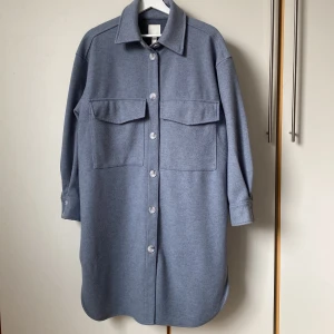 Duvblå lång overshirt från H&M - Lång duvblå overshirt från H&M. Den har långa ärmar och knappar framtill samt två stora bröstfickor. Dessutom två rymliga fickor i sidorna. Perfekt för lager-på-lager-stil under kyligare dagar. Längd 103 cm, mått runt midjan 130 cm. 