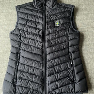 Svart dunväst från Cobolt of Sweden - - Snygg svart dunväst från Cobolt of Sweden med dragkedja och ett stilrent märke på bröstet. Vadderad och kviltad lättviktsväst i ett mjukt vindtätt och vattenavstötande yttertyg. Västen är vadderad med polyester med ”dunkänsla”. -Västen har en hög krage och två praktiska fickor med dragkedja. Ny pris ca 899kr! Vid frågor, tveka inte att höra av er 😊📦 (Storlek: 40/M, normal i storleken)