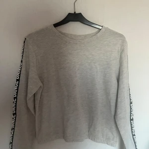 Grå långärmad tröja från KappAhl - Säljer en grå långärmad tröja från KappAhl med texten 'DANCE' längs ärmarna. Tröjan har en rund halsringning och är perfekt för en avslappnad stil. Passar bra till jeans eller leggings.
