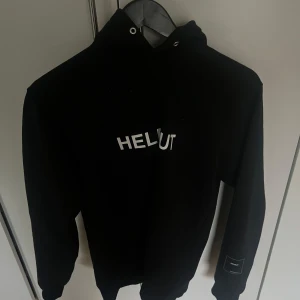 Svart hoodie från Helmut Lang - Säljer en stilren svart hoodie från Helmut Lang med tryck på bröstet och ärmen. Perfekt för en casual look. Den har en klassisk passform med långa ärmar och en bekväm huva. Skriv gärna om ni vill ha mera bilder eller om ni har någon fråga. 