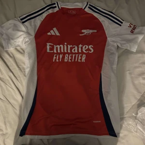 Arsenal hemmatröja 24/25 fotbollströja  - Säljer en snygg röd och vit fotbollströja från Adidas med korta ärmar. Tröjan har ett tryck av ett klubbemblem och sponsorlogga på framsidan. Perfekt för fotbollsfans!