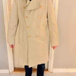 Filippa K trenchcoat med knappar - Snygg Filippa K trenchcoat med dubbelknäppning och klassisk krage. Jackan har långa ärmar och är perfekt för höst och vår. En stilren och tidlös design som passar till många tillfällen.