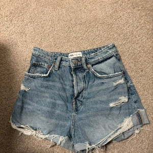 Blå jeansshorts från Zara - Säljer ett par snygga blå jeansshorts från Zara med fransiga kanter och slitna detaljer. Shortsen har en knappgylf och klassisk femficksdesign. Perfekta för en avslappnad stil! Säljer då de inte riktigt är min stil. Pris går alltid att diskutera och hör gärna av dig för fler frågor😊