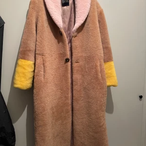 Fårpäls- mjuk och skön - Säljer en oanvänd mysig beige fårpälsjacka med gula ärmslut och en röd detalj på ryggen. Jackan har en rosa krage och knäpps med knappar framtill. Den är väldigt mjuk och i hög kvalitet. Foder i silke. 