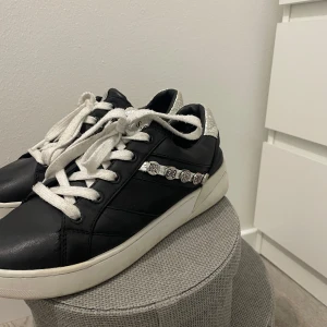 Guess skor - Snygga svarta sneakers från Guess med vita snören och silverdetaljer på sidan. Skorna har en vit sula och en glittrig häl med Guess-logga. Perfekta för en trendig look.