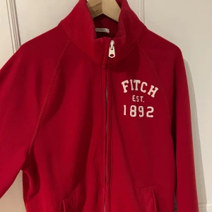 Röd hoodie från Abercrombie & Fitch - Säljer en snygg röd hoodie från Abercrombie & Fitch med dragkedja och texten 'FITCH EST. 1892' på bröstet. Perfekt för en avslappnad stil. Hoodien har långa ärmar och en hög krage.