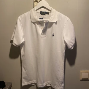 Vit pikétröja från Ralph Lauren - Säljer en klassisk vit pikétröja från Ralph Lauren med den ikoniska loggan broderad på bröstet. Tröjan har korta ärmar och knappar vid kragen. Perfekt för en stilren och avslappnad look.