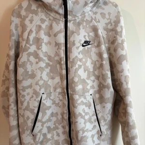 Nike tech camo - Säljer en snygg beige camouflagemönstrad hoodie från Nike. Den har en dragkedja framtill och två praktiska fickor. Perfekt för en avslappnad stil.