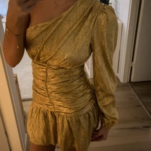 Guld one-shoulder klänning - Snygg Meet MeThere guldklänning med en axeldesign och långa ärmar. Den har en tight passform och veckade detaljer som ger den en stilren look. Perfekt för en festkväll! M/L liten storlek