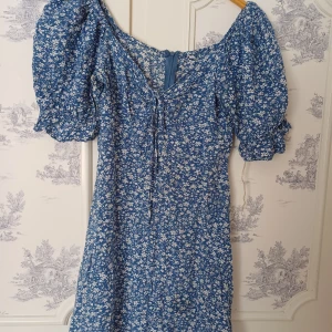 Blå blommig klänning med puffärmar - Charmig blå klänning med vita blommor och puffiga kortärmar. Den har en söt knytning framtill och en dragkedja bak. Perfekt för en somrig look!