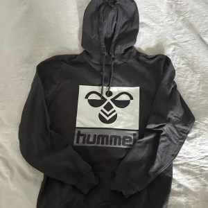 Svart hoodie från Hummel - Säljer en svart hoodie från Hummel med deras ikoniska logga på framsidan. Tröjan har en justerbar huva med snören och långa ärmar. Perfekt för en avslappnad stil eller till träning. Endast en liten defekt på ena snöret. 
