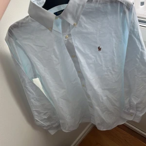 Vit skjorta från Ralph Lauren - Snygg vit skjorta från Ralph Lauren med klassisk krage och broderad logga på bröstet. Skjortan har långa ärmar och knappar framtill. Perfekt för en stilren look. Inte täcken på användhet!