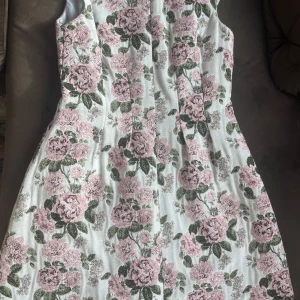 Blommig klänning med v-ringning - Säljer en vacker blommig klänning med v-ringning och ärmlös design. Klänningen har ett romantiskt mönster med rosa blommor och gröna blad på en vit bakgrund. Perfekt för sommarens alla tillfällen!