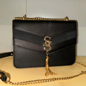 Svart axelväska med guldkedja - Snygg svart axelväska med guldkedja och dekorativ tofsdetalj. Väskan har ett elegant S-märke på framsidan. Den har en praktisk storlek för att bära dina nödvändigheter. Aldrig använd, som ny 