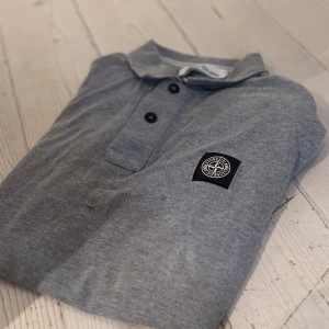 Grå pikétröja från Stone Island - Säljer en stilren grå pikétröja från Stone Island med klassisk krage och tre knappar framtill. Tröjan har en broderad logotyp på bröstet. Perfekt för en avslappnad men snygg look.