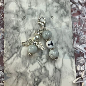 Ängelformad berlock med bokstav - Charmig ängelformad berlock med silverfärgade vingar och vita pärlor. En av pärlorna har bokstaven 'T'. Perfekt att fästa på ett armband eller halsband.